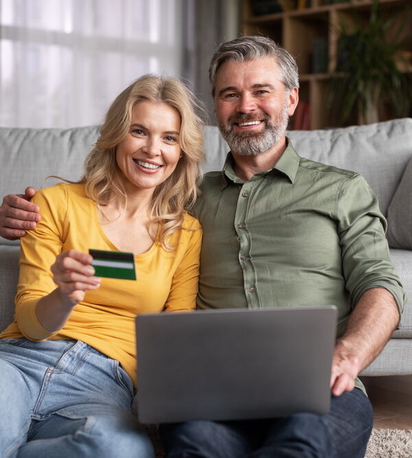 E-Commerce Concept. Smiling Middle Aged Husband And Wife With Laptop And Credit Card Making Online Shopping Or Virtual Payments While Relaxing At Home Together, Happy Spouses Smiling At Camera