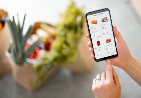 Holding phone with running e-shop application and bags full of fresh food on the background. Shopping groceries online concept