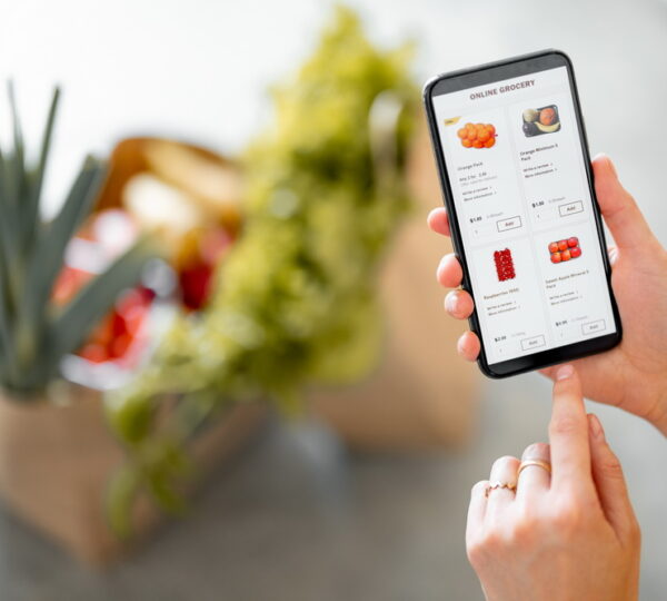 Holding phone with running e-shop application and bags full of fresh food on the background. Shopping groceries online concept