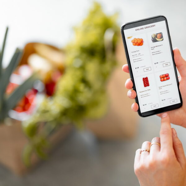 Holding phone with running e-shop application and bags full of fresh food on the background. Shopping groceries online concept