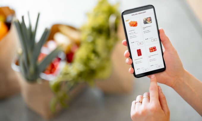 Shopping groceries online Holding phone with running e-shop application and bags full of fresh food on the background. Shopping groceries online concept